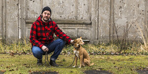 Mann mit karierter Jacke kniend vor einer Friedhofsmauer mit seinem braunen Hund