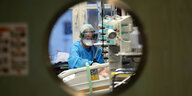 Blick durch ein rundes Fenster in eine Intensivstation: Man sieht eine Pflegerin in Schutzausrüstung an einem Patient:innenbett