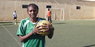 Eine Spielerin steht auf einem Fußballplatz und hält einen Ball in der Hand. Hinter ihr trainieren zwei Menschen, dahinter eine Mauer