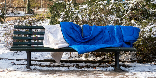 Auf einer Bank nahe eines verschneiten Parks liegt ein blaue Schlafsack