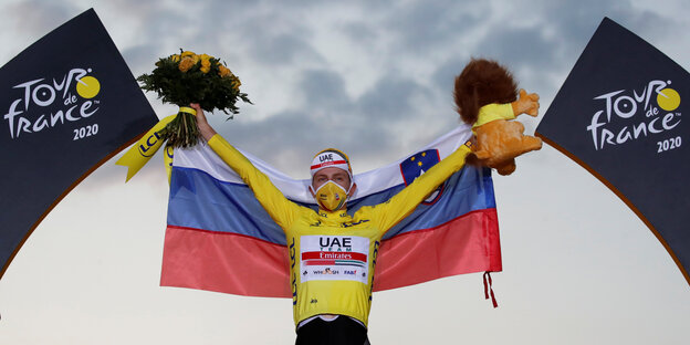 Der Radler Tadej Pogacar jubelt bei der Siegerehruzng der Tour de France mit einem Blumenstrauß in der einen und einem Plüschlöwen in der anderen Hand