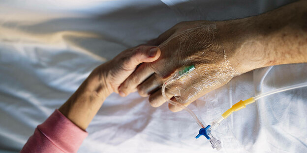 Eine Frauenhand hält eine ältere Hand eines Patienten in der eine Infusion gelegt ist