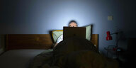An Mann liegt im Bett und schaut auf den Bildschirm seines Laptops