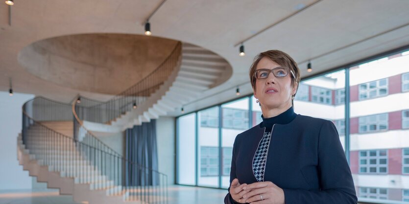 Eine Frau im blauen Kostüm und mit Brille steht im hellen, lichten Treppenhaus des Deutschlandhauses