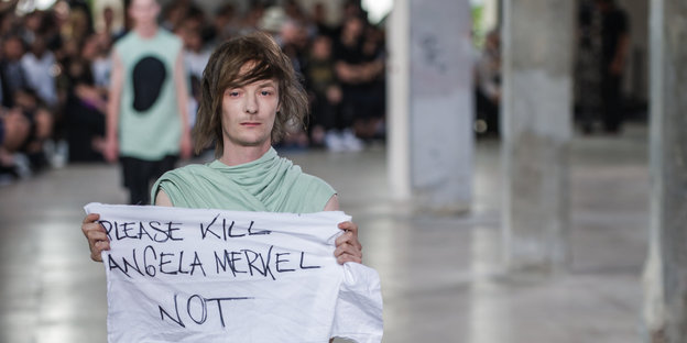 Ein männliches Modell auf dem laufsteg einer Modenschau hölt ein Tuch, auf dem „Please Kill Angela Merkel – Not“ steht