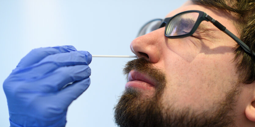 Eine behandschuhte Hand steckt einem bärtigen Mann mit Brille ein Wattestäbchen in die Nase