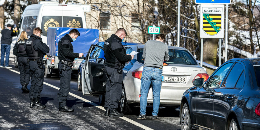 Eine Schlange von Autos ist zu sehen, davor stehen Männer. Rechts das sächsische Wappen und das Grenzschild der Bundesrepublik