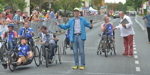 Eine Gruppe von Handbikern und ein stehender Mann mit geöffneten Armen, links hinter einer Absperrung Zuschauer