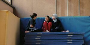Drei Mädchen, eins davon trägt Kopftuch, sitzen auf einem Matten-Berg in einer Sporthalle