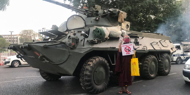 Ein buddhistischer Mönch protestiert vor einem Panzerfahrzeug mit einem Schild in der Hand, das ein durchgestrichenes Maschinengeweher zeigt
