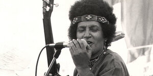 Eine Schwarze Frau mit einem Afro, darin ein Stirnband. Sie singt in ein Mikrofon und hält eine Gitarre