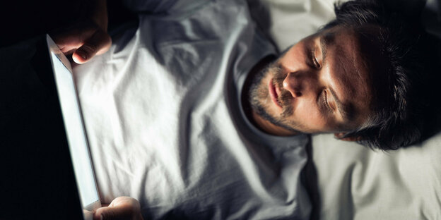 Ein Mann liegt im Bett und sieht sich etwas auf einem Tablet an Ein Mann liegt im Bett und sieht sich etwas auf einem Tablet an