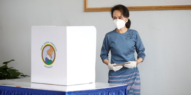 Aung San Suu Kyi, die Regierungschefin von Myanmar schmeißt ihre Stimme in die Wahlurne. Sie ist eine alte Frau und trägt Mund-Nasen-Schutz und eiße Handschuhe.
