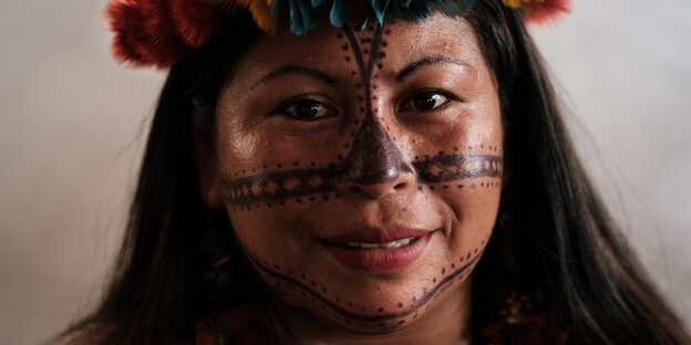 Alessandra Munduruku, ene Frau mit glatten dunklen, langen Haaren und dunklen Augen. Sie trägt Kopfschmuck. In der Mitte ihrer Stirn die Nase hinab und von einer Wange quer zur anderen ist sie bemalt.