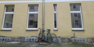 Eine sanierte Häuserwand mit mehreren Fenstern und einer Regenrinne, an die ein Fahrrad angeschlossen ist. Unten bricht die Fassade ab und es ist die Bausubstanz zu sehen.