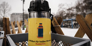 Eine gelbe Pumpkanne steht in einem grauen Kasten. Auf der Kanne steht in schwarzer Schrift "Auffüllort für heißes Wasser" und mit roter Schrift "Kostenlos". Im Hintergrund sieht man einen Zaun und Schnee.