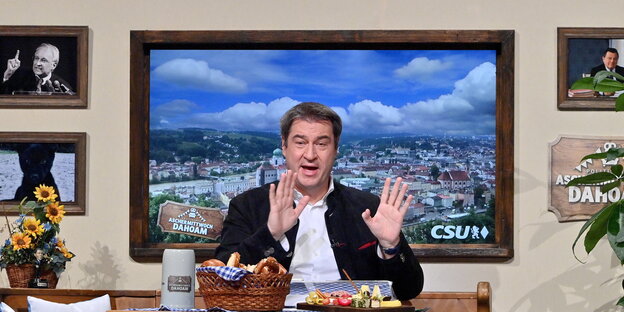 Markus Söder sitzt beim ins Digitale verlegten Politischen Aschermittwoch in einer Art Wohnzimmer. Auf einer Eichenbank, vor ihm ein Tisch mit weiß-blauer Decke, Bierkrug und deftiger Brotzeit - an der Wand hinter ihm hängt ein Bild von Passau