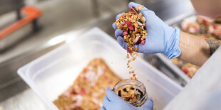 Eine Hand mit blauem Gummihandschuh füllt Müsli in ein Glas ab