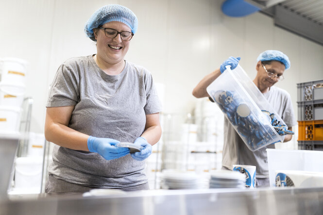 Zwei Menschen mit Haarschutz und Handschuhen in einer Art Großküche