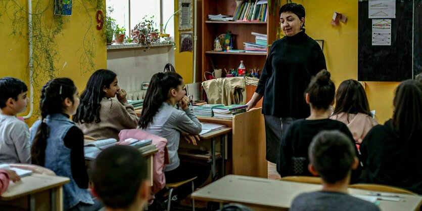 Kinder sitzen in Schulbänken. Im Hintergrund eine Lehrerinn