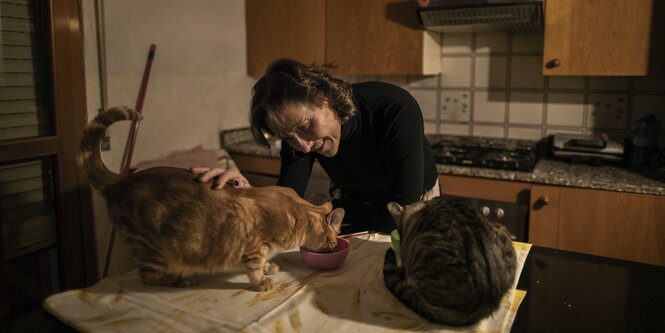 Renata Colombi zuhause in ihrer Küche, vor ihr auf dem Tisch sitzen zwei Katzen