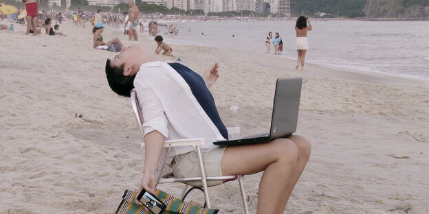 Auf einem Klappstuhl am Strand sitzt ein junger Mensch, Smartphone in der Hand, Laptaop auf den Knien.