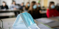 Eine hellblaue OP-Maske liegt auf einem Tisch, im Hintergund sind Schüler:innen an Tischen zu sehen