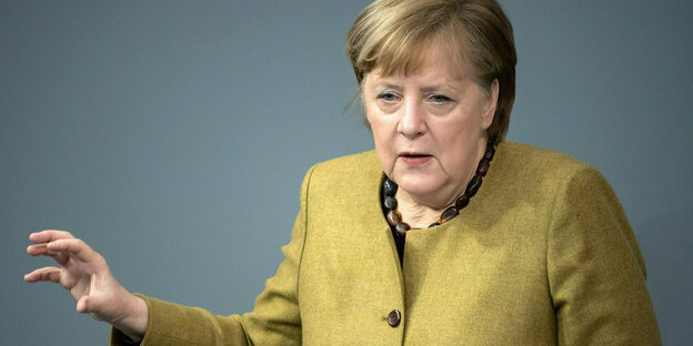 Angela Merkel steht in einem senfgelben Blazer an einem Redepult und gestikuliert.
