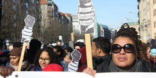 Eine Frau hält den oberen Rand eines Transparents. Dahinter Schilder mit der Aufschrift "Wir fordern ein Denkmal"