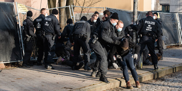 eine große Gruppe Polizisten vor einem eingedrückten Bauzaun, zwei drängen eine Person ab