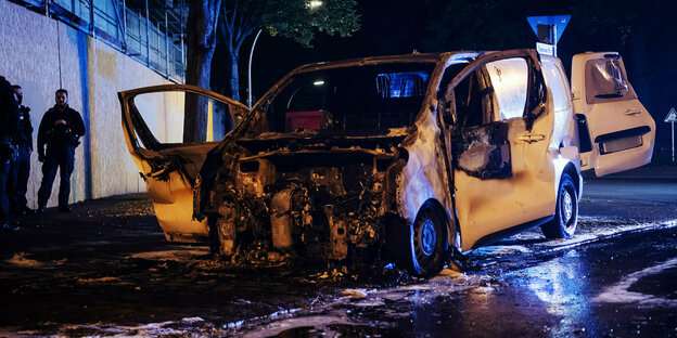 Ein abgebranntes Auto steht nachts an der Straße. Zwei Polizisten stehen daneben.