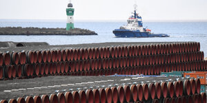 Das Bild zeigt gestapelte Rohre an einer Küste, im Hintergrund ein Schiff und einen Leuchtturm.