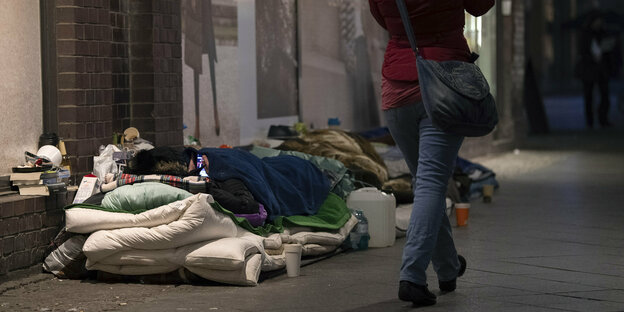 Obdachloser liegt unter vielen Decken auf einer Matratze in der Berliner Friedrichstraße. Fußgängerin geht vorbei