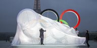 Mann zieht Plane von überdimensional großen Olympiaringen herunter, im Hintergrund Eifelturm