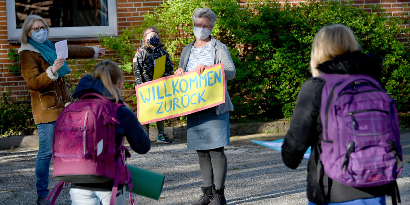 Eine Frau hält ein Schhild "Willkommen zurück"; zwei Kinder mit Ranzen laufen auf sie zu