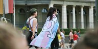 Zwei Frauen laufen auf einer Straße, eine von ihnen trägt eine Fahne mit einem feministischen Symbol ls Umhang