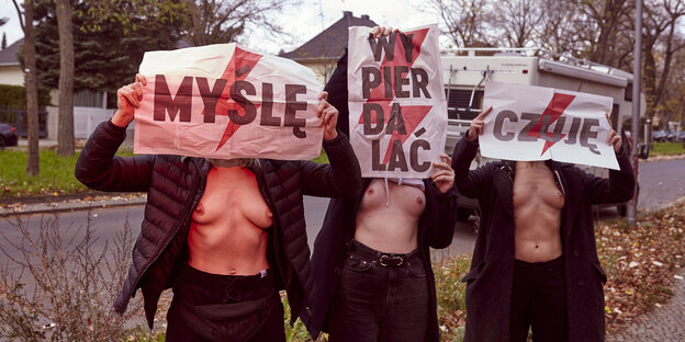 Drei Frauen mit freiem Oberkörper und Plakate mit Slogans vor dem Gesicht