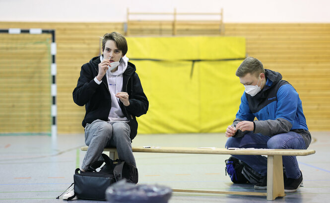 Zwei Schüler sitzen in einer Turnhalle und bohren mit Stäbchen in der Nase