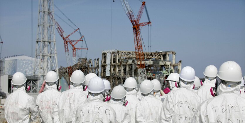 Menschen Schutzanzügen stehen vor den zerstörten Reaktorgebäuden des AKW Fukushima circa ein Jahr nach der Katastrophe