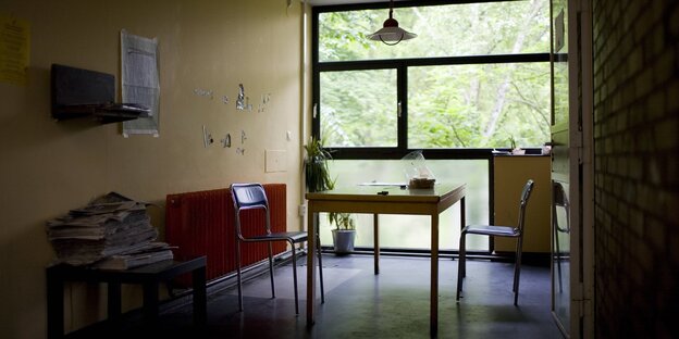 Innenansicht eines Zimmers im Studentendorf Schlachtensee: Ein Tisch, zwei Stühle, ein Stapel Zeitungen. An den Wänden hängen noch Fetzen von hier vorher platzierten Postern oder Plakaten.