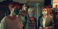 Drei Frauen und ein Mann, alle tragen Masken, stehen in einem Raum, der ein Wohnzimmer sein könnte