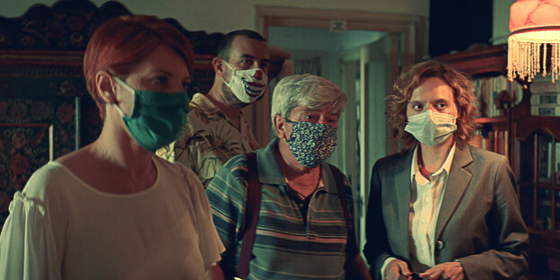Drei Frauen und ein Mann, alle tragen Masken, stehen in einem Raum, der ein Wohnzimmer sein könnte
