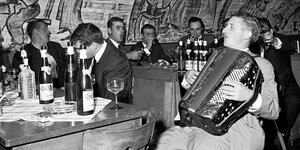 Männer sitzen in einem Kellergewölbe und trinken Wein, singen, einer spielt Akkordeon