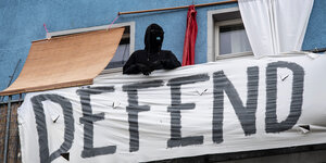 Ein Vermummter steht auf einem Balkon, an dem ein "Defend"-Banner hängt