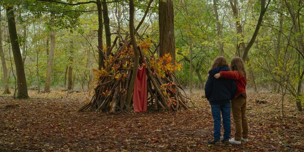 Zwei Mädchen stehen mit dem Rücken zum Betrachter und schauen auf eine selbstgebaute Hütte aus Ästen im Wald