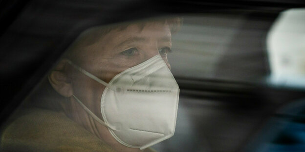 Angela Merkel etwas verdunkelt hinter einer Autofensterscheibe, sie blickt besorgt und trägt eine FFP2-Maske