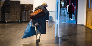 Ein Mensch trägt einen blauen Beutel. Über dem Arm hängt eine weitere Tasche. Er geht auf braunem Boden auf eine geöffnete Tür zu.