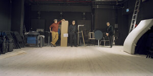 Die drei Musiker stehen in einem Aufnahmestudio, schwarze Wände, Holfußboden, kahler Raum, an der Wand lehnen Klappstühle