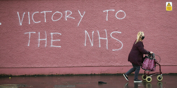 Frau mit Rollator läuft an einer roten Mauer vorbei, auf der steht: Victory to the NHS