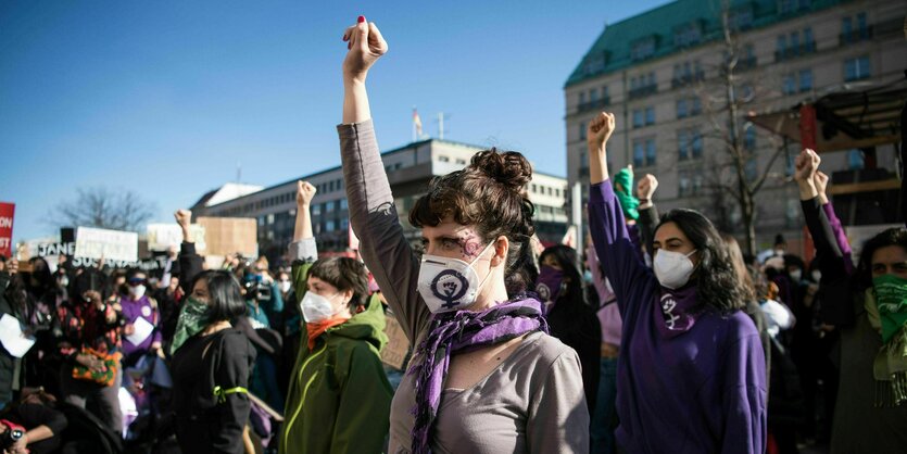 Menschen mit Masken stehen in einer Straße und recken die rechten Finger zur Faust. Im Hintergrund sieht man einen blauen Himmel ohne Wolken.